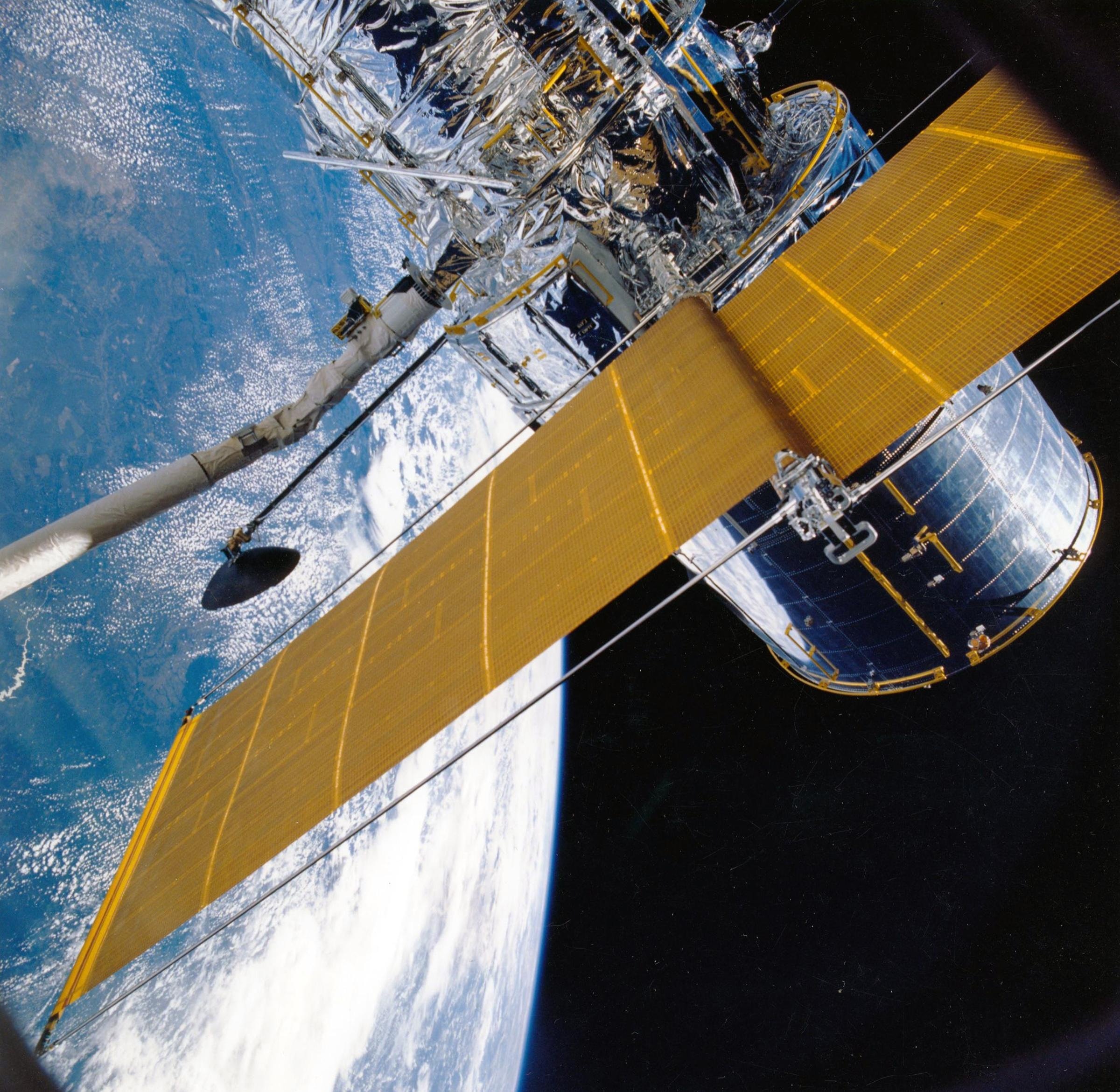 Partial view of a satellite in space. Pictured is part of the hull, a solar panel array and the Earth in the background.